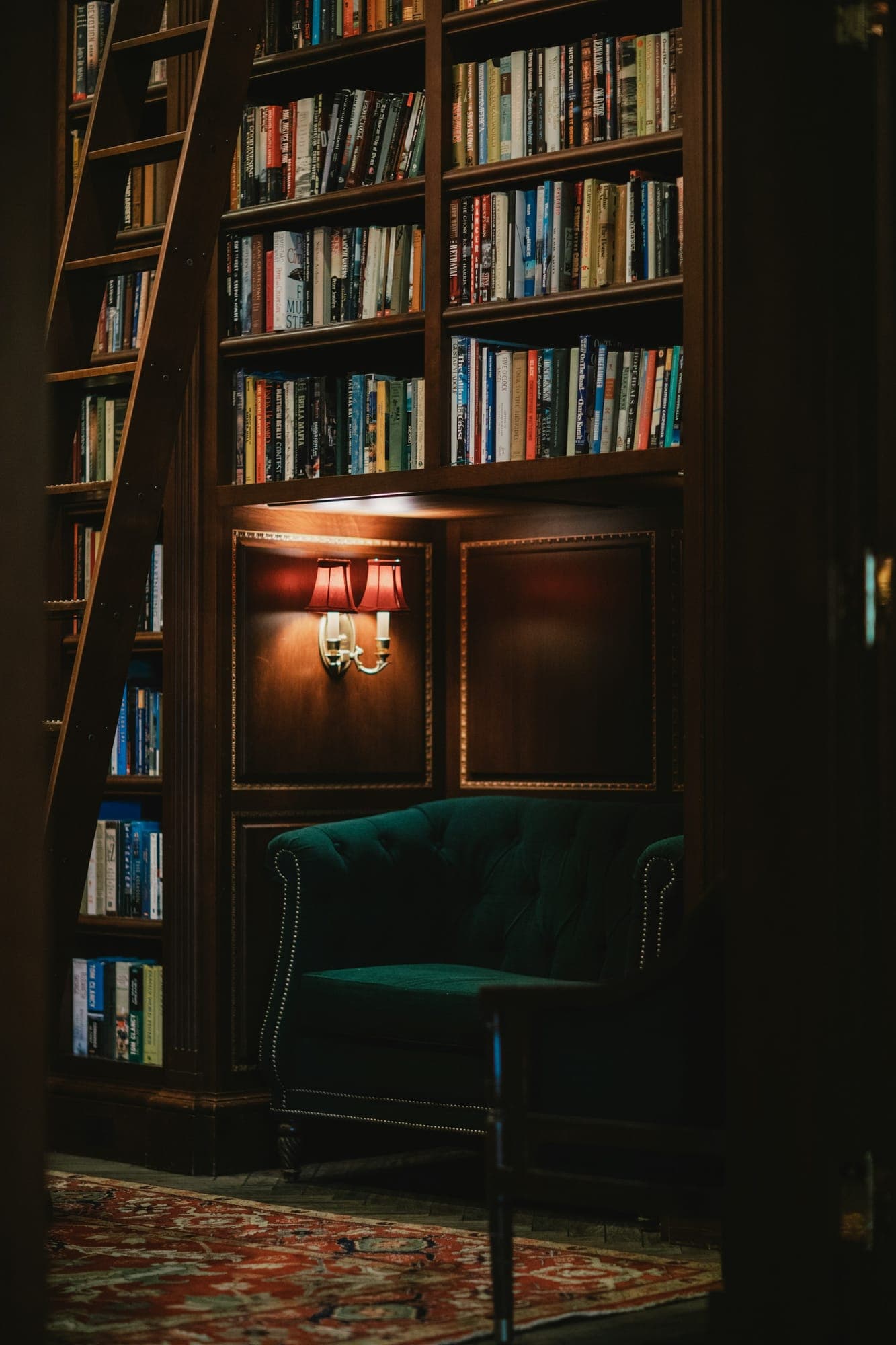 A dark paneled library with a rolling ladder, sconces, and a velvet chair